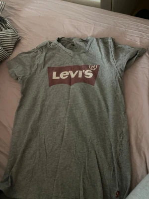Grå Levi's t-shirt med logga - Säljer en klassisk grå t-shirt från Levi's i bomull med den ikoniska röda och vita loggan framtill. T-shirten har korta ärmar och normal passform, perfekt till jeans eller shorts. Enkel och stilren design som funkar till det mesta.