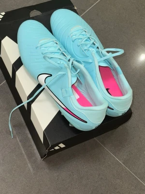Nike blå fotbollsskor med dobbar - Snygga ljusblå fotbollsskor från Nike med klassisk vit och svart swoosh på sidan. Skorna har snörning, rosa innersula och är tillverkade i syntetmaterial. Perfekta för dig som vill sticka ut på planen med stilrena och moderna skor.