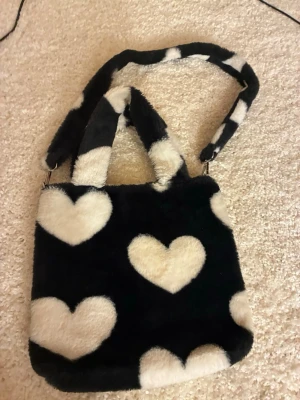 Fluffig svart handväska med hjärtan - Säljer en fluffig svart handväska med vita hjärtan i fuskpäls. Väskan har två handtag och en avtagbar axelrem i samma mönster. Invändigt finns ett fack med dragkedja. Perfekt för dig som gillar gulliga accessoarer och vill sticka ut.
