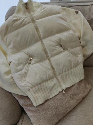 Vit dunjacka från Moncler - Snygg vit dunjacka från Moncler med quiltad front och rygg, ribbade muddar och hög krage. Jackan har två dragkedjeförsedda fickor fram och stor broderad Moncler-logga på ryggen. 