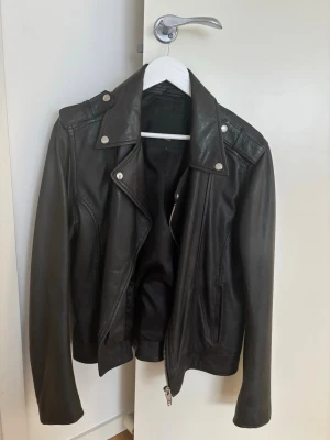 Svart klassisk skinnjacka rockanblue  - Snygg svart skinnjacka i bikerstil med silvriga detaljer och dragkedja framtill. Jackan har bred krage, axelklaffar och två fickor med dragkedja. Perfekt för dig som vill ha en tidlös och cool look.