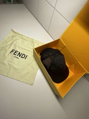 Svart keps från Fendi med logomönster - Säljer en svart keps från Fendi med klassiskt FF-logomönster. Kepsen har mesh-paneler på sidorna för extra ventilation och justerbar rem bak. Kommer med original dustbag och gul Fendi-box. Perfekt för dig som gillar exklusiva accessoarer.