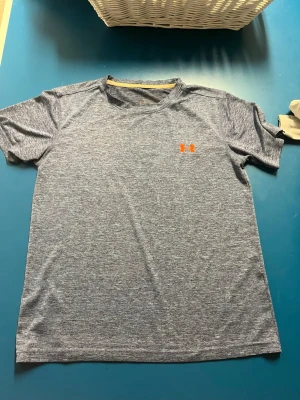  Under Armour T-shirt  - Snygg tränings-tshirt från Under Armour med orange logga på bröstet. Tillverkad i ett lätt och snabbtorkande funktionsmaterial som passar perfekt till gymmet eller löprundan. Klassisk passform med korta ärmar och rund hals.
