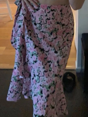 Rosa blommig omlottkjol - Säljer en lång omlottkjol i rosa med blommigt mönster. Kjolen har volangdetaljer och knyts i midjan för en snygg och bekväm passform. 