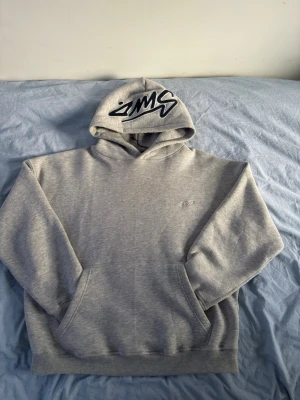 Grå Pull&Bear hoodie med broderad logga - Snygg grå hoodie från Pull&Bear med stor broderad logga i svart och vitt på huvan och en mindre logga på bröstet. Klassisk känguruficka framtill och mjukt material som känns skönt mot huden. Perfekt för en avslappnad streetwear-look.
