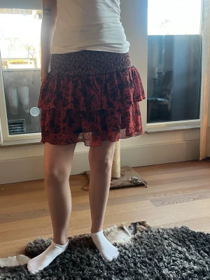 Röd blommig volangkjol - Kort röd kjol med svart blommönster och dubbla volanger. Kjolen har en smockad midja för skön passform och är gjord i ett lätt, luftigt material som ger en flowy känsla. Perfekt för dig som gillar en feminin och trendig stil.