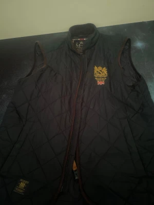 Svart quiltad dunväst från Morris - Snygg svart dunväst från Morris med quiltat mönster och guldfärgad logga på bröstet. Västen har bruna detaljer längs dragkedjan och ärmarna samt brittisk flagga på insidan. Perfekt för lager-på-lager och streetstyle.
