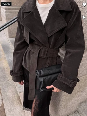 Mörkbrun trenchcoat i mocka - Stilren mörkbrun trenchcoat i mjuk mocka med breda slag, knytskärp i midjan och spännen vid ärmsluten. Klassisk och oversized passform som ger en cool look. Perfekt för dig som vill ha en trendig och tidlös rock.