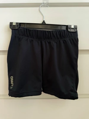 Svarta träningsshorts från Craft - Snygga svarta träningsshorts från Craft med elastisk midja och diskret logga på vänster ben. Tillverkade i ett lätt och snabbtorkande material, perfekta för löpning eller gym. Bakficka med dragkedja för småsaker. Sportig och enkel design.