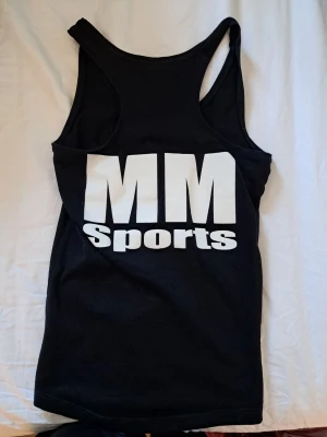 Svart linne från MM Sports - Säljer ett svart träningslinne från MM Sports med stort vitt tryck på bröstet. Linnet har brottarrygg och är gjort i ett mjukt material som passar perfekt för gymmet eller annan träning.