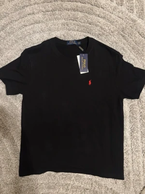 Svart t-shirt från Polo Ralph Lauren - Svart t-shirt från Polo Ralph Lauren med klassisk rund hals och den ikoniska röda logotypen broderad på bröstet. Tillverkad i mjuk bomull som känns skön mot huden. Enkel och stilren design som passar till det mesta.