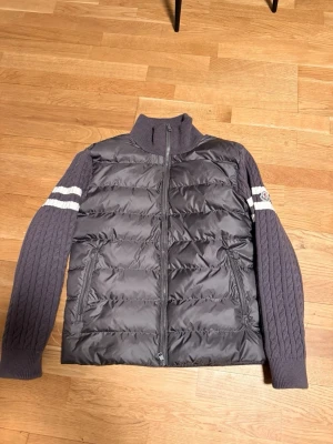 Grå Moncler cardigan  - Helt ny moncler cardigan oanvänd Snygg grå dunväst från Moncler med stickade, kabelmönstrade ärmar och vita ränder. Västen har hög krage, dragkedja framtill och två fickor. Moncler-logga på vänster ärm. Perfekt för kyliga dagar och riktigt trendig look.