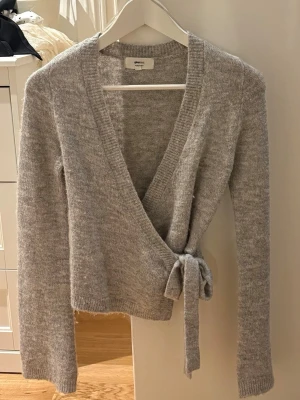 Grå omlottkofta från Gina  - Mysig grå omlottkofta från ginamed knytband i midjan och långa ärmar. Koftan är stickad i mjukt material och har en djup v-ringning. Perfekt för lager på lager och enkel att styla till olika outfits.