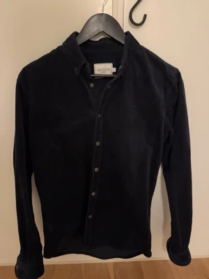 Svart manchesterskjorta Les Deux - Stilren svart skjorta från Les Deux i mjuk manchester. Klassisk krage, knappar framtill och långa ärmar med knapp vid manschetten. Perfekt för dig som gillar en enkel men snygg look. Storlek s men passar mer som m