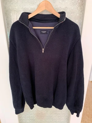 Marinblå halvzip tröja från Holebrook - Säljer en marinblå stickad tröja från Holebrook med halvzip och hög krage. Tröjan har ett snyggt strukturmönster och en röd logopatch på ärmen. Perfekt för dig som gillar klassisk svensk design och vill ha något både stilrent och bekvämt. XL i storlek men passar även L!