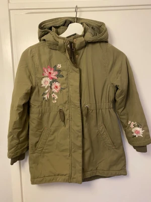 Olivgrön jacka med blommig brodyr - Stilren olivgrön jacka med huva och dragsko i midjan. Jackan har snygga broderade blommor i rosa och vitt på bröstet och ena ärmen. Två snedställda fickor framtill och muddar vid ärmsluten. Perfekt för kyliga dagar och ger en trendig look.