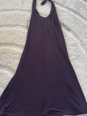 Svart långklänning med halterneck - En enkel och stilren svart långklänning med halterneck och lös passform. Klänningen har en mjuk och följsam känsla, perfekt för varma dagar eller kvällar. Inga synliga detaljer eller mönster, bara klassisk och tidlös design.