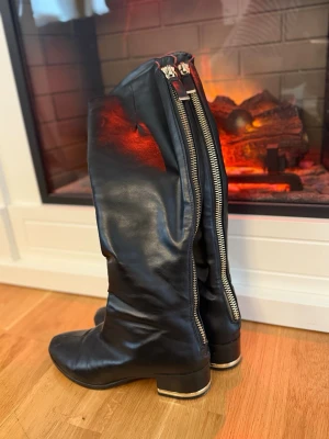 Svarta höga läderstövlar med dragkedja - Säljer ett par svarta höga stövlar i mjukt läder med grov silverfärgad dragkedja längs baksidan. Stövlarna har låg klack med metallisk detalj och rundad tå. Perfekta för dig som vill ha en edgy och stilren look.