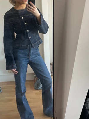 Mörkblå jeansjacka med silverknappar - Säljer en mörkblå jeansjacka med cool asymmetrisk knäppning och silverfärgade knappar. Jackan har markerade sömmar och en figurnära passform med lite utsvängda ärmar. Perfekt för dig som gillar en unik och trendig look.