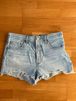 Ljusblå Levi's 501  jeansshorts med fransar storlek W31 - Säljer ett par ljusblå jeansshorts från Levi's 501 med råa, fransiga kanter och klassisk femficksdesign. Shortsen har hög midja och är tillverkade i bomull. Perfekta för sommaren och ger en avslappnad vibe. Storlek W31