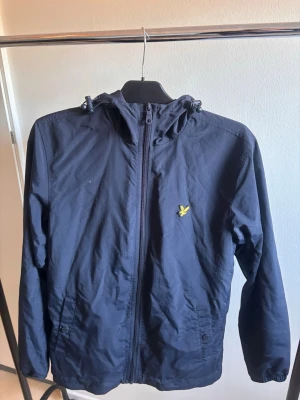 Marinblå vindjacka från Lyle & Scott - Snygg marinblå vindjacka från Lyle & Scott med gul logga på bröstet. Jackan har huva med dragsko, hel dragkedja framtill och två sidofickor. Tillverkad i lätt och vindtätt material, perfekt för blåsiga dagar. Använd nån enstaka gång, storlek S.