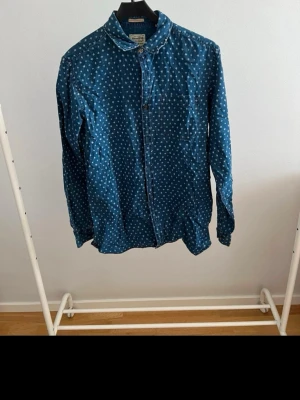 Blå prickig skjorta med knappar - Snygg blå skjorta med vita prickar och klassisk krage. Skjortan har långa ärmar och knäppning framtill. Perfekt för dig som vill ha en unik och trendig look. Materialet känns mjukt och skönt mot huden.