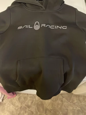 Svart hoodie från Sail Racing storlek 160 - Svart hoodie från Sail Racing med stor logga framtill. Klassisk känguruficka och huva. Tillverkad i mjukt och bekvämt material, perfekt för chill dagar eller när du vill ha en sportig look. Snygg och enkel design som funkar till det mesta.