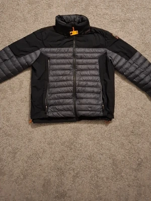 Svart och grå pufferjacka Parajumpers  - Snygg svart och grå pufferjacka från Parajumpers med hög krage och dragkedja framtill. Jackan har quiltade partier, två sidofickor med dragkedja och en patch med PJS-logga på ärmen. Perfekt för kalla vinterdagar.