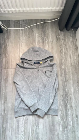 Grå zip hoodie från Polo Ralph Lauren - Snygg grå hoodie med dragkedja från Polo Ralph Lauren. Klassisk design med huva, ribbade muddar och det ikoniska broderade logotypen på bröstet. Perfekt för en chill och stilren look. Tillverkad i mjukt bomullsmaterial. Detaljerade frågor, fråga gärna på 😁 Storlek: S OBS! Kommer med snöret, vill man det!