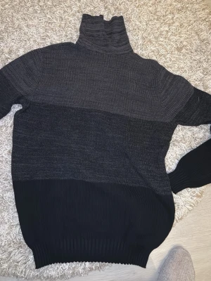 Mörkgrå stickad polotröja - Stickad polotröja i olika nyanser av mörkgrått och svart. Tröjan har breda färgblock, hög polokrage och ribbstickade muddar vid ärmslut och nederkant. Perfekt för kalla dagar och ger en stilren look.