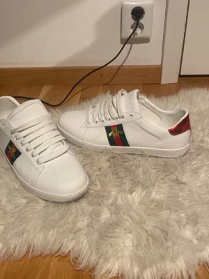Gucci vita sneakers med röd detalj - Säljer ett par vita Gucci sneakers med klassiska gröna och röda ränder på sidan samt en guldbroderad bi. Baksidan har metallicdetaljer i rött och svart med Gucci-loggan. Skorna är låga och har vita snören. Perfekta för dig som vill sticka ut med lyxig streetstyle. Skorna är lite slitna på insidan av hälen, samt att 3-4 stygn har släppt längst ner vid snörerna på ena skon! 