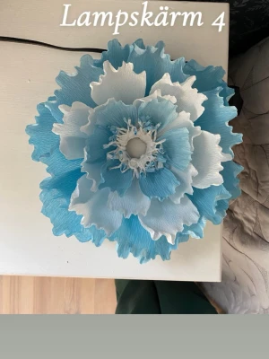 Bordslampa i form av en blomma  - Endast lampskärm säljs! Egentillverkad!  Höjd: ca 12 cm  Bredd: ca 25 cm 1. Lampan tål inte direkt solljus, då kommer färgen med störst sannolikhet att blekas tyvärr. Men den är ej passande för t ex fönsterbrädan. Den passar bäst vid ett nattduksbord, mörkare rum där solen ej når fram så mycket, eller t ex en byrå mitt i ett rum. (Var extra försiktig under sommaren!!) så tråkigt när den bleks. Jag rekommenderar att köpa en svagaste glödlamporna då den är gjord av crepepapper.