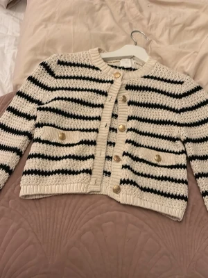 Randig stickad kofta med guldknappar - Snygg stickad kofta i vitt och svart med horisontella ränder och guldfärgade knappar framtill. Två dekorativa fickor med knappar på framsidan. Långärmad och ribbad vid ärmslut och nederkant. Perfekt för en trendig och avslappnad look.