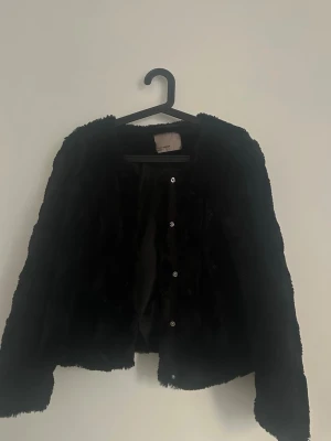 Svart pälsjacka från Vero Moda - Snygg svart pälsjacka från Vero Moda med mjuk fuskpäls och knäppning framtill. Jackan har rundad halsringning och är helsvart både på utsidan och insidan. Perfekt till våren 💞