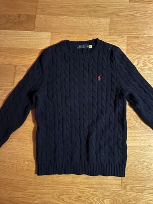 Mörkblå kabelstickad tröja Polo Ralph Lauren - Kabelstickad mörkblå tröja från Polo Ralph Lauren med rund halsringning och röd broderad logga på bröstet. Tröjan har långa ärmar och ribbade muddar vid ärmslut och nederkant. Perfekt för lager på lager och klassisk stil.