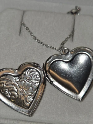 Halsband med hjärtformat medaljong - Snyggt halsband i silverfärgat stål med hjärtformat medaljonghänge. Medaljongen har ett vackert graverat blommönster på framsidan och kan öppnas för att lägga in ett foto eller minne. Kedjan är tunn och är i rostfritt stål 45 cm. Perfekt accessoar för en personlig touch.