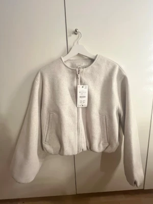  cropped cardiganjacka Zara - Trendig ljusgrå cardiganjacka från Zara med rund halsringning och dragkedja framtill. Jackan har två fickor, ballongärmar och en croppad passform som ger en modern look. Tillverkad i mjukt material som känns skönt mot huden.
