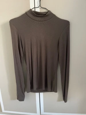 Brun ribbad polotopp - Säljer en brun ribbad polotopp med lång ärm. Toppen har en hög krage och är figurnära i modellen, perfekt att styla med jeans eller kjol. Materialet är mjukt och stretchigt för en bekväm känsla.