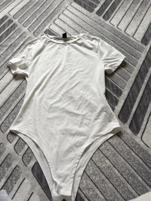 Vit kortärmad body - Säljer en enkel och stilren vit body med korta ärmar och rund hals. Bodyn har en klassisk passform och är tillverkad i ett mjukt, stretchigt material som sitter bekvämt mot kroppen. Perfekt att matcha med jeans eller kjol.