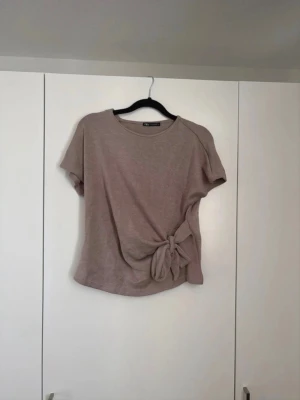 Beige topp med knyt från Zara - Säljer en beige topp från Zara med korta ärmar och rund halsringning. Toppen har en snygg knytdetalj i sidan som ger en trendig look. Perfekt att matcha med jeans eller kjol för en avslappnad stil.