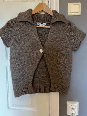 Brun stickad kortärmad kofta Zara - Kortärmad stickad kofta från Zara i en mjuk brun nyans. Koftan har en bred krage, en stor vit knapp framtill och ribbstickade kanter.