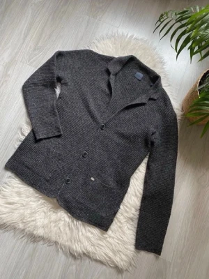 Mörkgrå ribbad kofta med knappar - Skön mörkgrå kofta med ribbad struktur och tre knappar framtill. Koftan har två fickor och en klassisk krage. Perfekt att slänga över en t-shirt för en chill look. Hör av er om frågor!