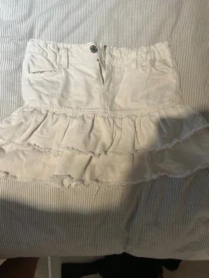 Vit jeanskjol från Gina Tricot - Säljer en vit jeanskjol från Gina Tricot med volangdetaljer och råa kanter. Kjolen har fickor fram, bälteshällor och dragkedja med knapp framtill. 