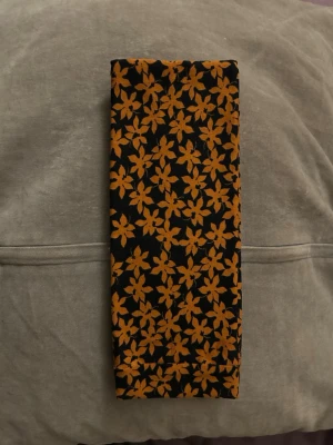 Svart hårband med orange blommönster - Snygg hårband med tätt orange blommönster. Tunn och mjuk, perfekt att knyta runt halsen eller i håret för en färgklick till din outfit. Mönstret ger en retrovibe och sticker verkligen ut.