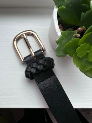 Svart skärp från H&M, 95cm - Svart skärp från H&M med en elegant guldfärgad spänne. Skärpet har en flätad detalj nära spännet som ger det en unik touch. Perfekt för att ge din outfit det lilla extra. Storlek: 95x2,5cm (XS)