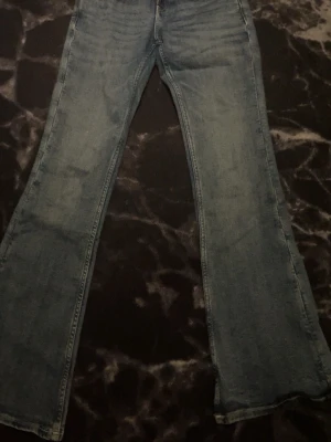 Blå bootcut jeans med fickdetaljer - Säljer ett par blå bootcut jeans med snygga fickdetaljer bak. Jeansen har klassisk femficksdesign, lätt slitna partier och utsvängda ben. Perfekta för dig som gillar retrostil och vill ha ett par jeans som sticker ut lite extra.
