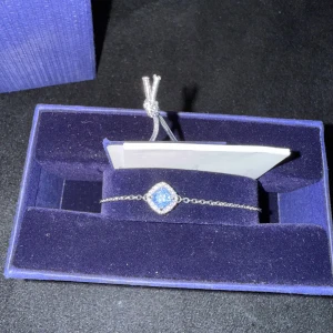 Gnistrande armband  - Ett elegant armband från Swarovski med tunn kedja i silverfärg och en fyrkantig, gnistrande ljusblå kristall i mitten, omgiven av små klara stenar. Justerbar längd med snygg dragsko. Perfekt för dig som gillar stilrena smycken med lite extra bling. 