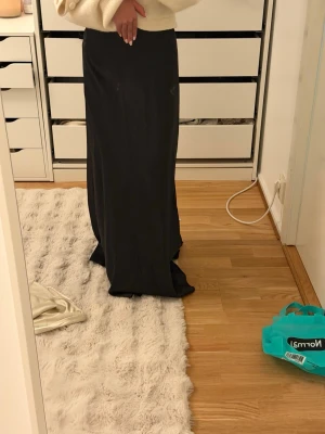 Svart långkjol i satin - Säljer en svart långkjol i glansigt satinmaterial. Kjolen har en rak och elegant passform som faller snyggt längs benen. Perfekt för dig som gillar en stilren och klassisk look. Helt ny aldrig använd storlek xxs