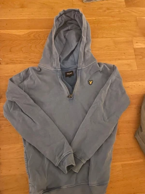Blå hoodie från Lyle & Scott - Snygg ljusblå hoodie från Lyle & Scott med halv dragkedja och klassisk gul logga på bröstet. Hoodien har huva och långa ärmar, tillverkad i mjukt bomullsmaterial. Perfekt för en avslappnad och stilren look.