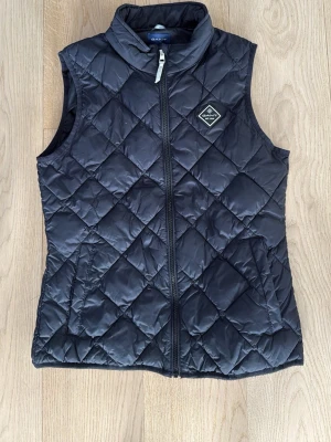 Svart quiltad dunväst från GANT - Svart dunväst från GANT med quiltat mönster och hög krage. Västen har dragkedja framtill och två sidofickor. GANT-logga på bröstet. Perfekt för lager-på-lager och ger en clean look.